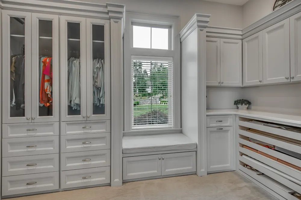 A gray painted mudroom with a window seat and glass cabinets by Imperial Cabinets