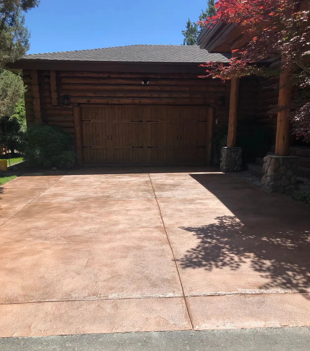 A color-sealed concrete driveway in front of a log home garage with pine trees