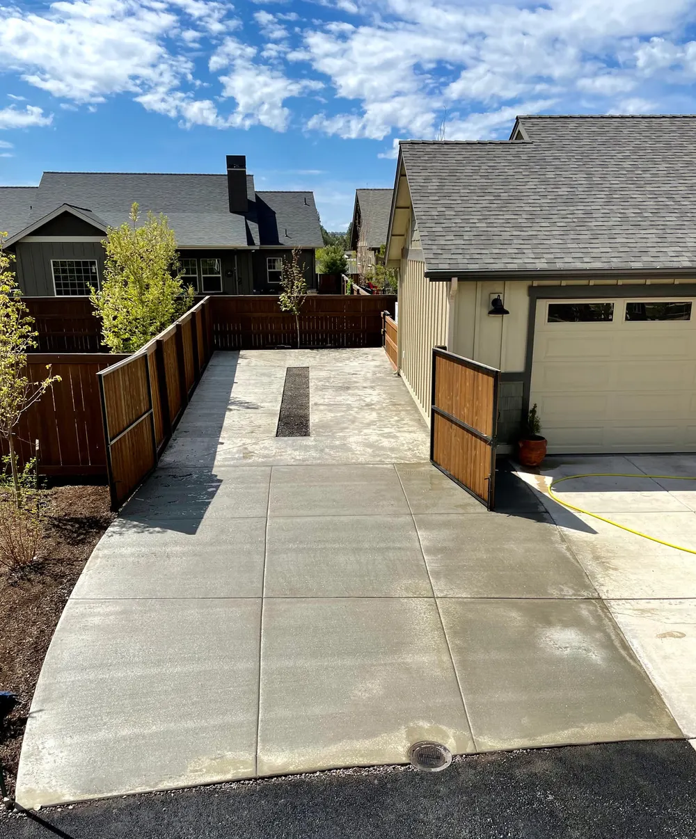 A new concrete side-yard drive with saw-cut joints leading through a wood gate past a garage