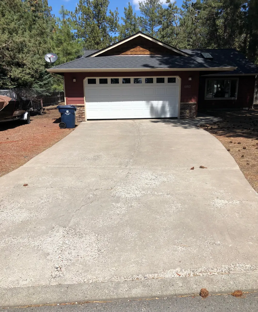 A smooth concrete driveway curving up to a white garage door, with pine needles and red cinder landscaping