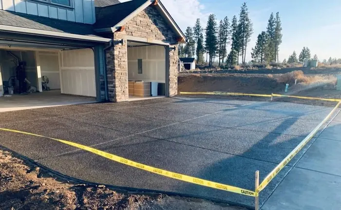 A new exposed-aggregate concrete driveway taped off in front of a stone-trimmed garage, with pines behind