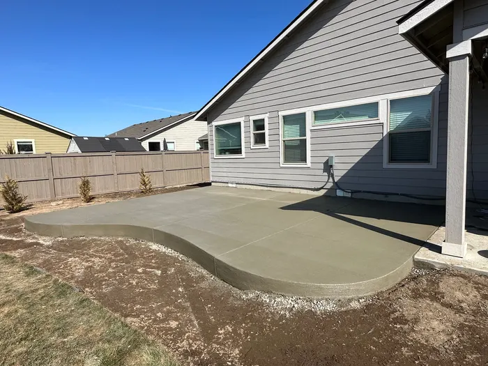 A new curved broom-finish concrete patio behind a gray house with a wood fence