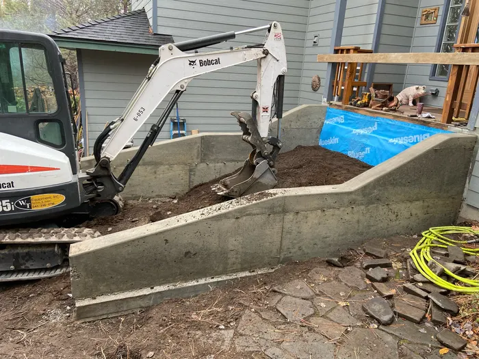 A new poured concrete retaining wall stepping down a yard beside a house, with a mini excavator