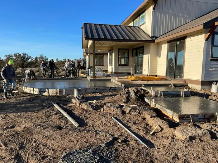 A crew finishing a fresh concrete patio slab behind a new high-desert home