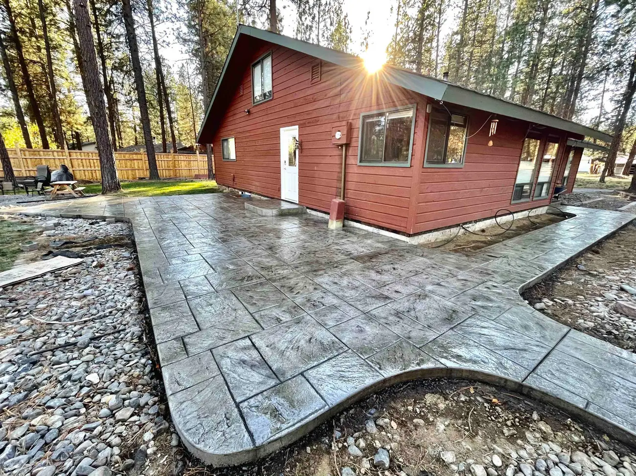 A new gray stamped-concrete patio wrapping a red house under tall ponderosa pines in Sisters, Oregon