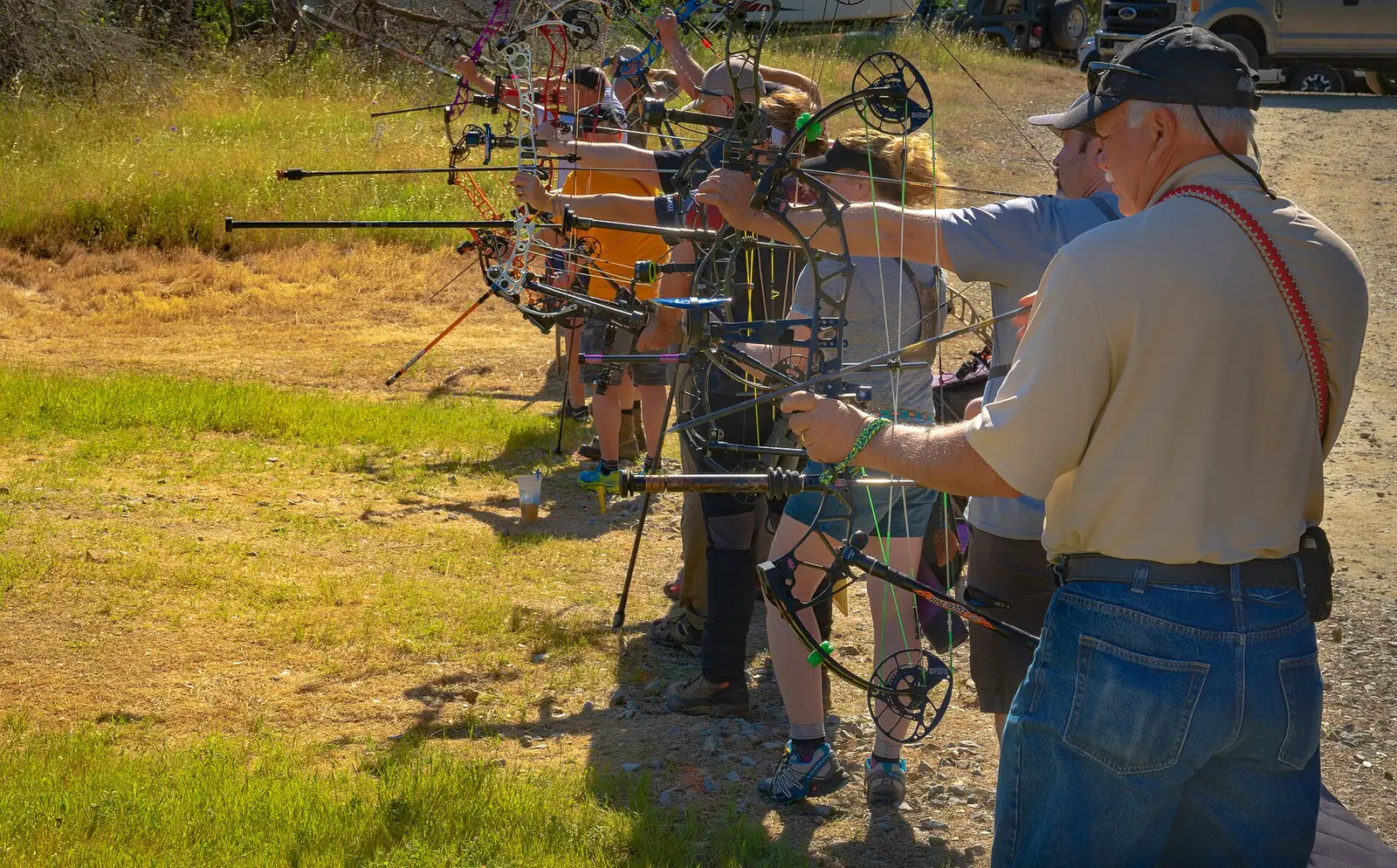 A line of archers at full draw with compound bows on an outdoor range, an older man in a cap watching in the foreground