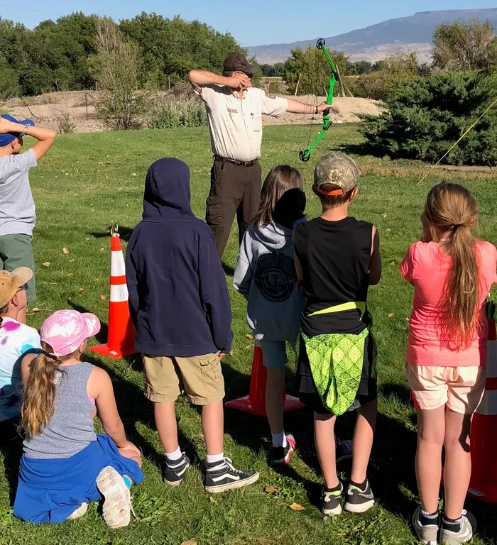 An instructor holds a green youth compound bow at full draw while a group of fourth-graders watches on a grassy field