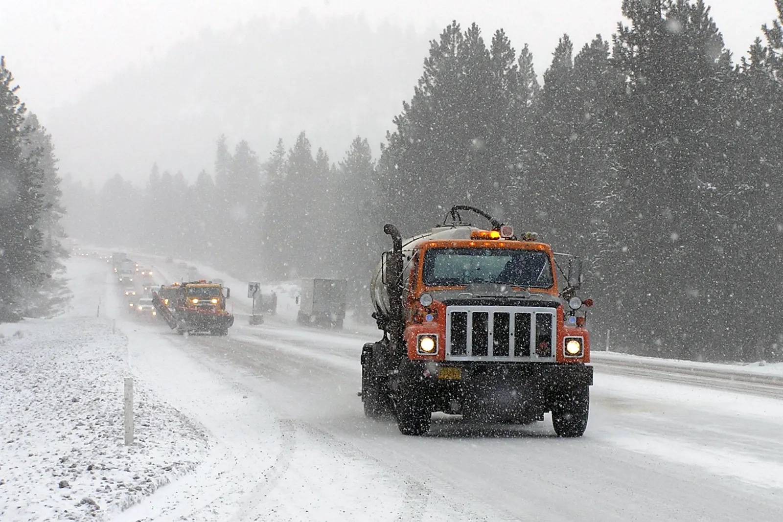 An ODOT tanker truck sprays liquid deicer on snowy U.S. 97 south of Bend, with a plow and traffic behind it among ponderosa pines