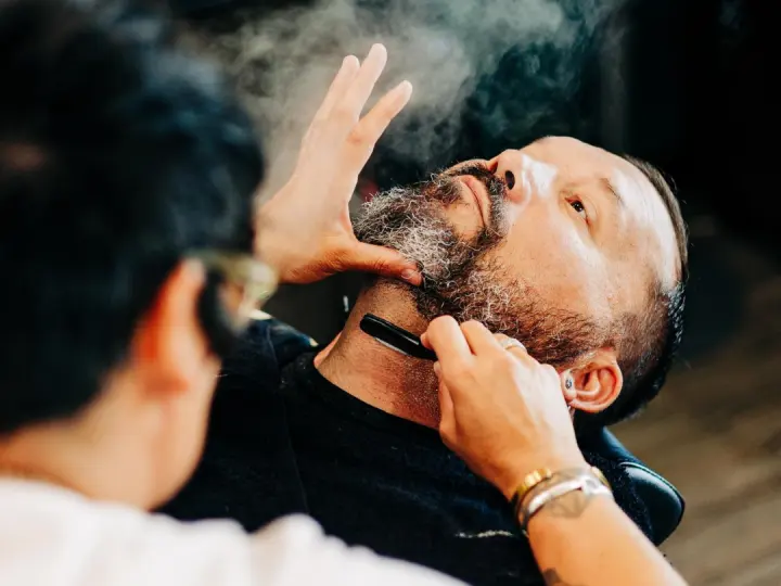 A barber shaping a client’s beard with a straight razor at The Roost in Bend