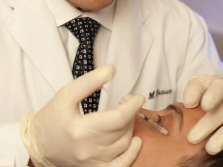A physician in a white coat and gloves injecting a patient’s forehead