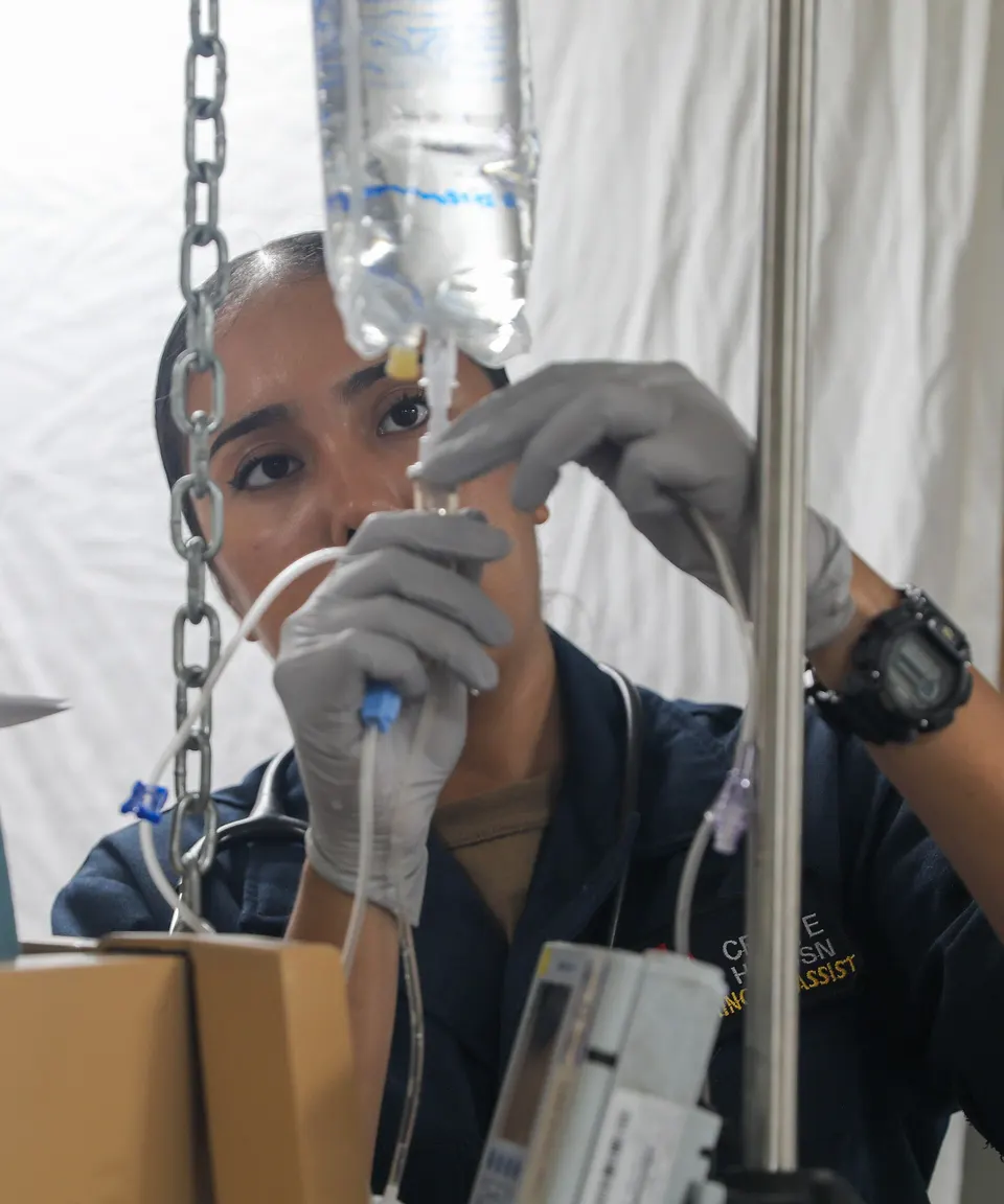 A gloved medical worker adjusting an IV bag and tubing on a pole