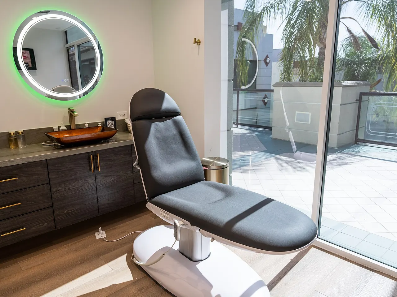 A bright med spa treatment room with a reclining treatment chair, wood cabinets and a lit round mirror