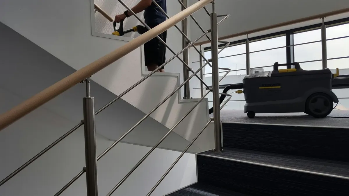 A technician cleans carpeted stairs with a hand tool connected to a portable carpet extraction machine on the landing
