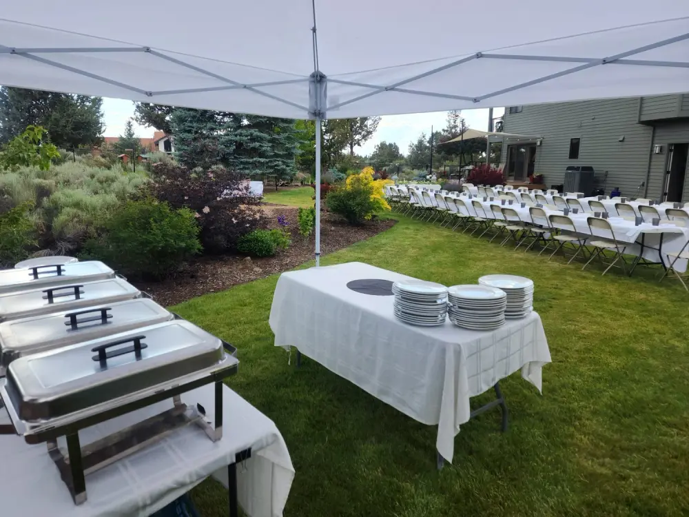 Chafing dishes and plate stacks under a pop-up tent on a Bend backyard lawn, with rows of chairs and sagebrush beds
