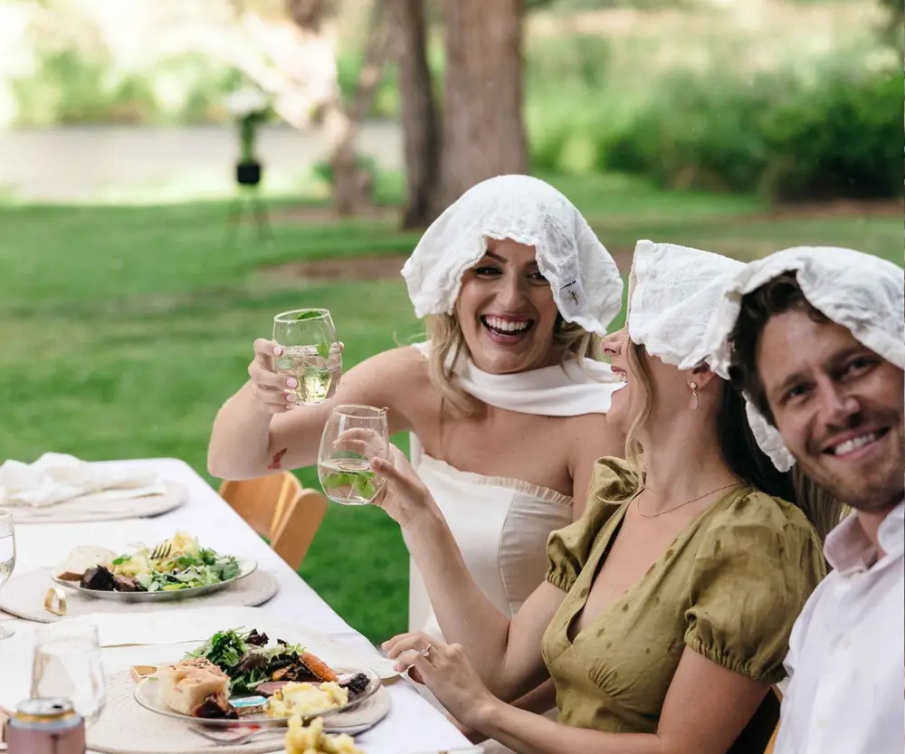 A bride and guests laughing and toasting at a long outdoor wedding table on a riverside lawn, catered by Bleu Bite