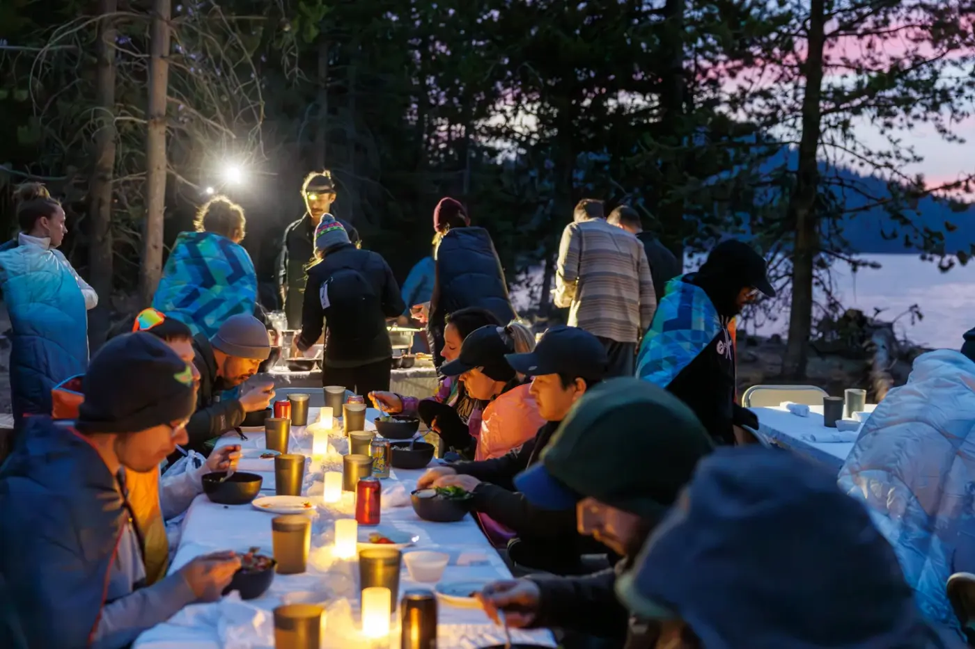 Guests in puffy jackets at a long candlelit table under the pines at dusk beside Paulina Lake, served by AlpenGlow Adventure Catering