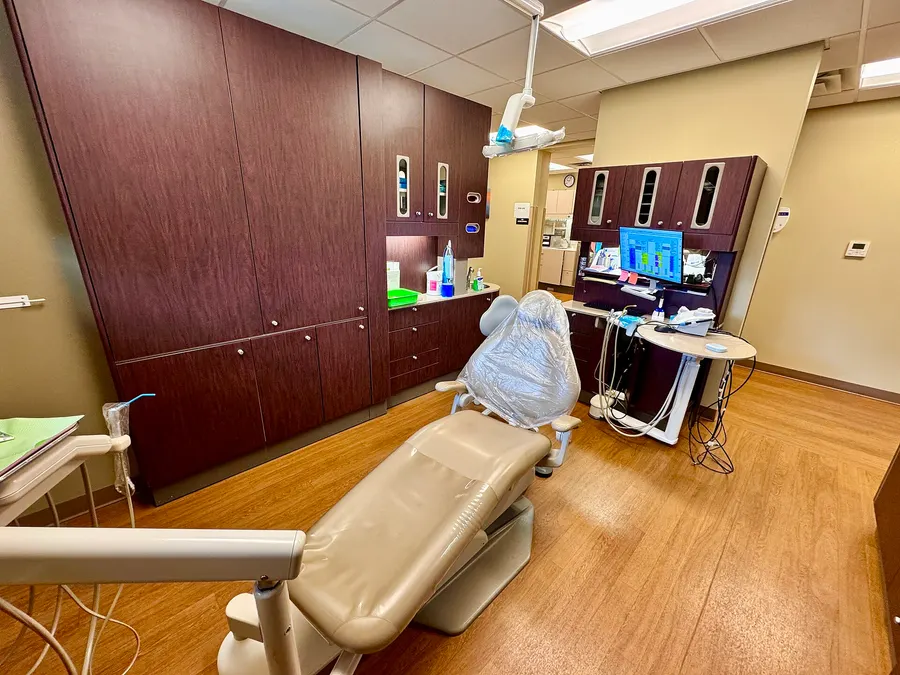 A dental exam room with a beige patient chair, overhead light, cabinets and a monitor showing a schedule