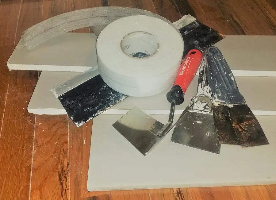 Pieces of half-inch drywall with a roll of mesh tape, a utility knife and several taping knives