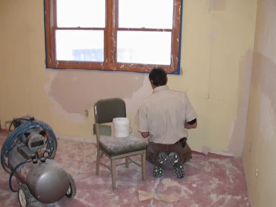 A man kneeling to patch walls in a room with masked windows, a compressor and a bucket of compound