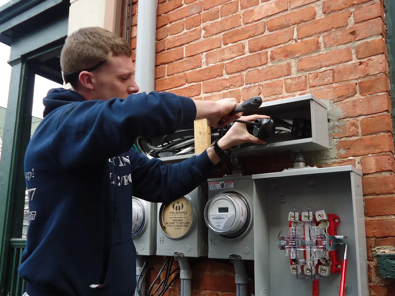 An electrician working on an outdoor meter base and service panels mounted on a brick wall