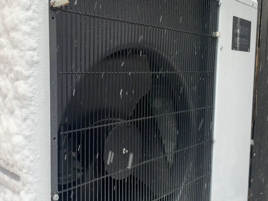 The fan grille of an air-source heat pump outdoor unit running with snow on the ground around it