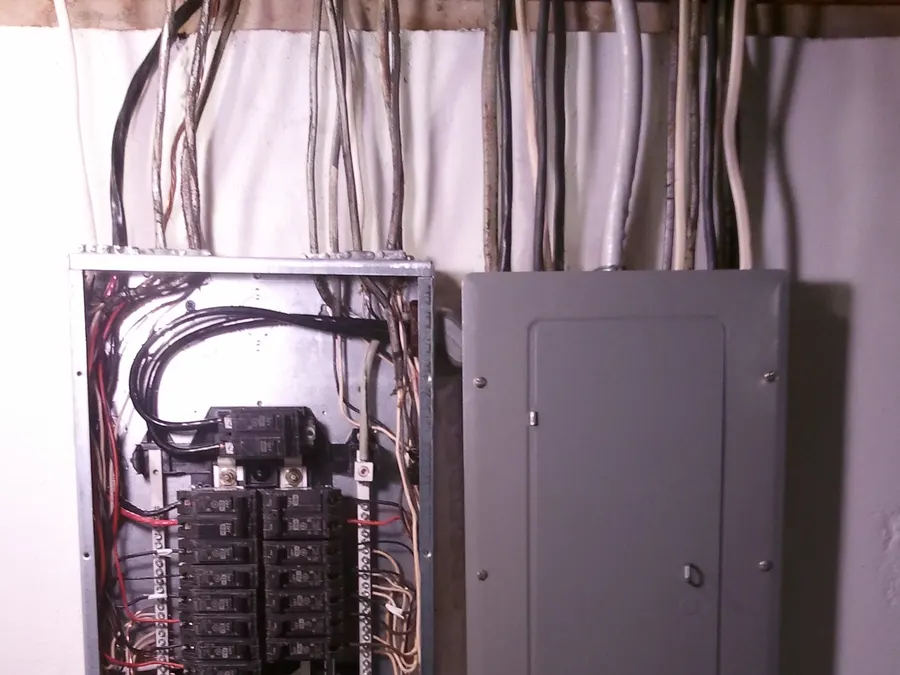 A gray main electrical panel beside a subpanel with its cover removed, showing breakers and wiring