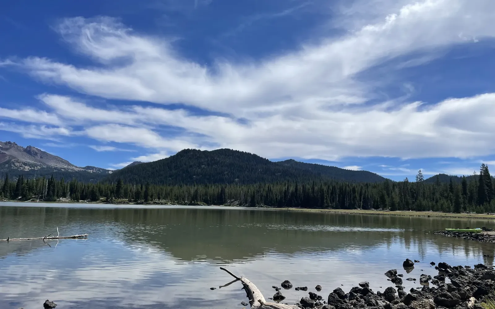 Sparks Lake west of Bend on a summer day, with calm water, lava rock and a bleached log in the foreground and forested buttes under white clouds