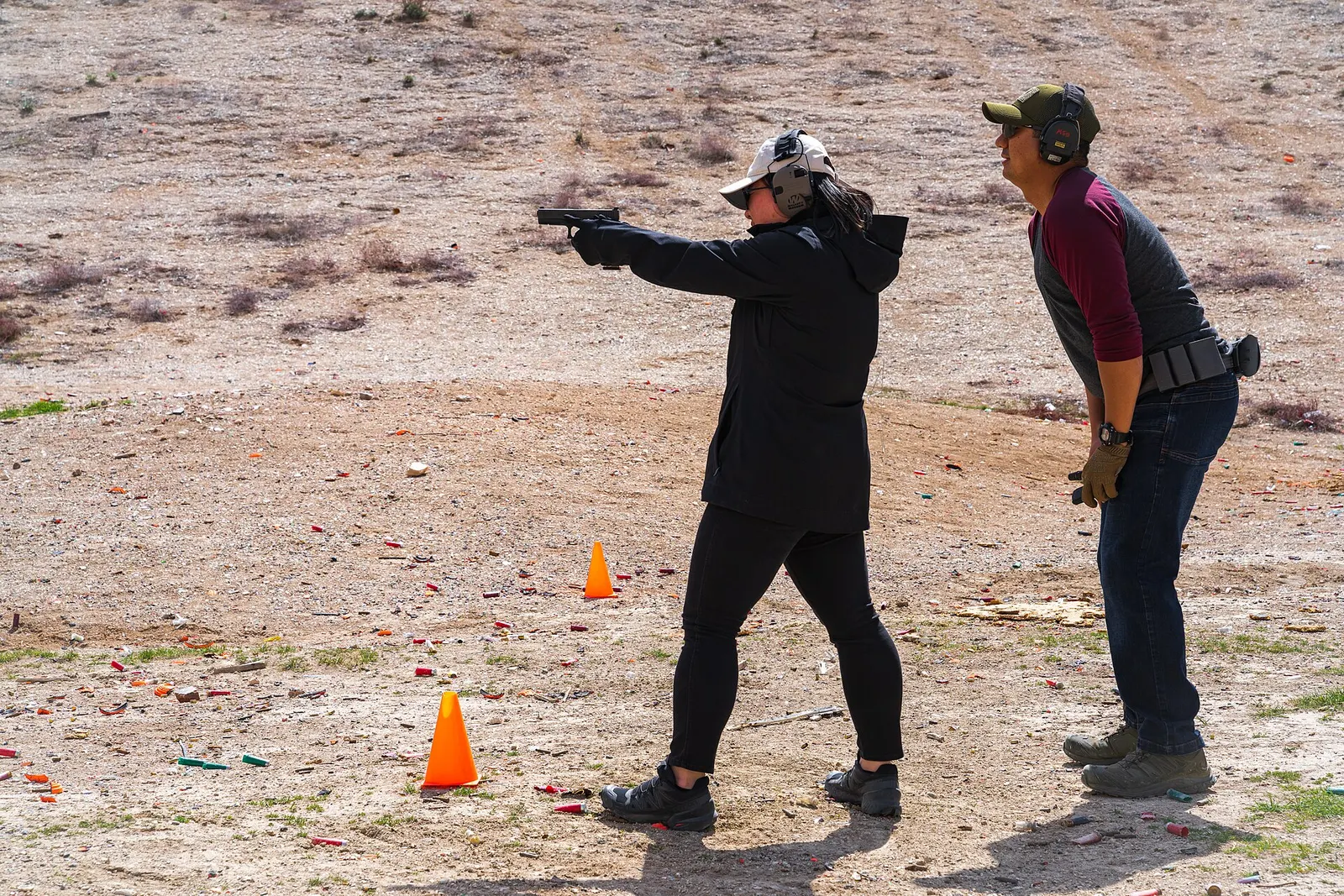 A range instructor coaches a new shooter firing a pistol at an outdoor dirt range, both wearing hearing and eye protection