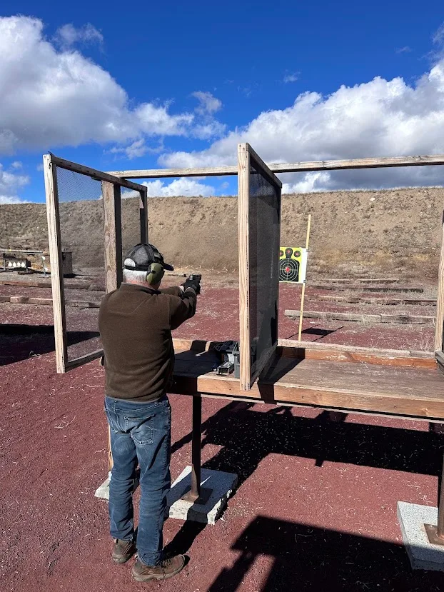 A shooter in ear protection fires a pistol from a wooden bench bay at Redmond Rod & Gun Club, with a target and red cinder berm downrange