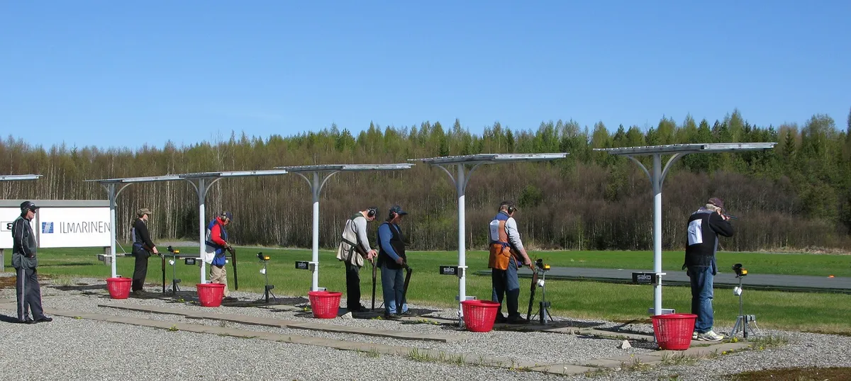 Shooters standing at stations along an outdoor trap shooting range with a tree line behind