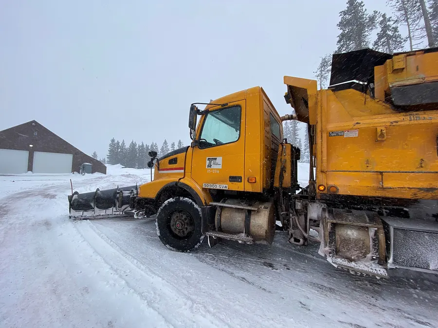 An orange ODOT snowplow with a wing blade parked on a snowy road beside pines in Bend