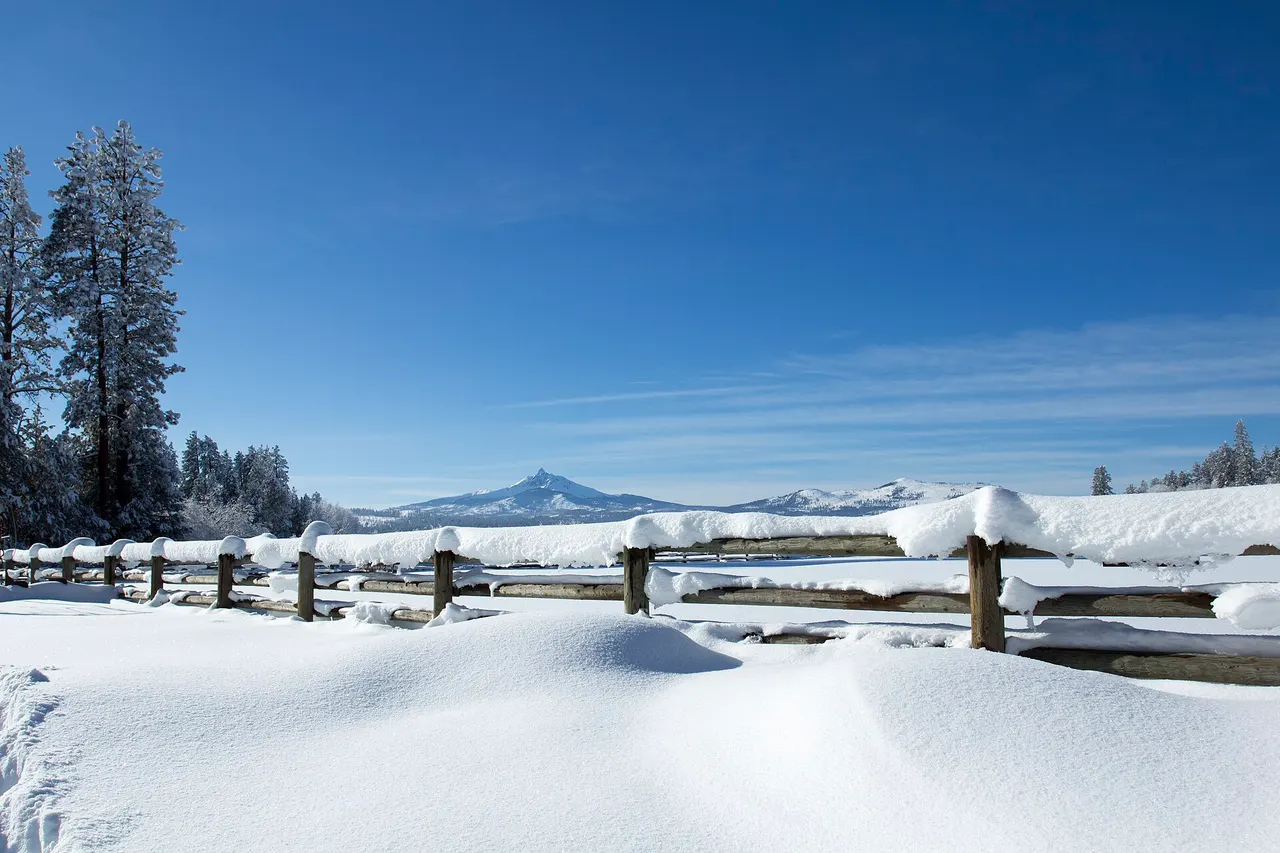 Deep snow drifted over a split-rail fence at Black Butte Ranch with Mount Washington under a clear blue sky