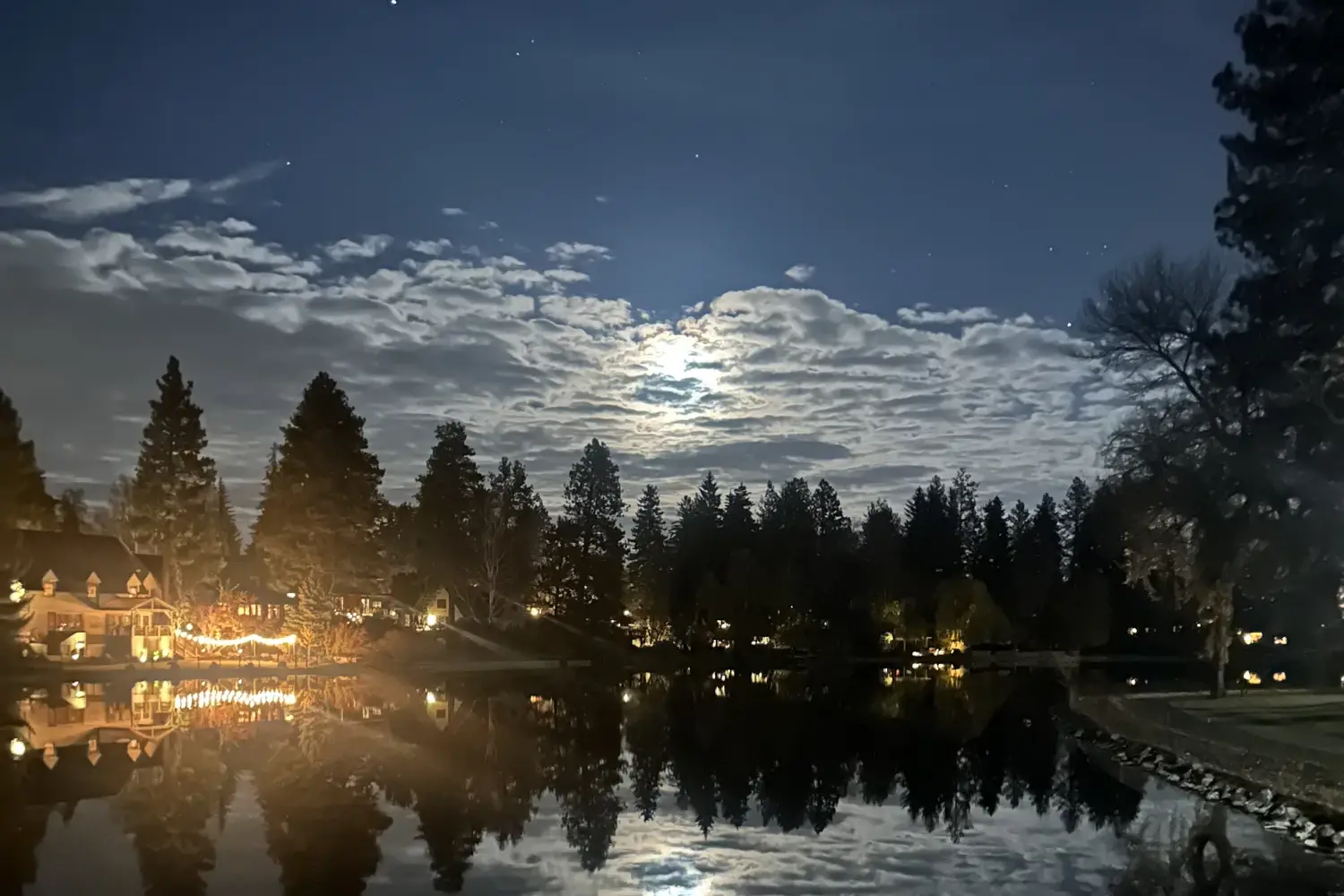A full moon glowing through clouds over Mirror Pond at night, with dark pines and warm lights reflected in the water