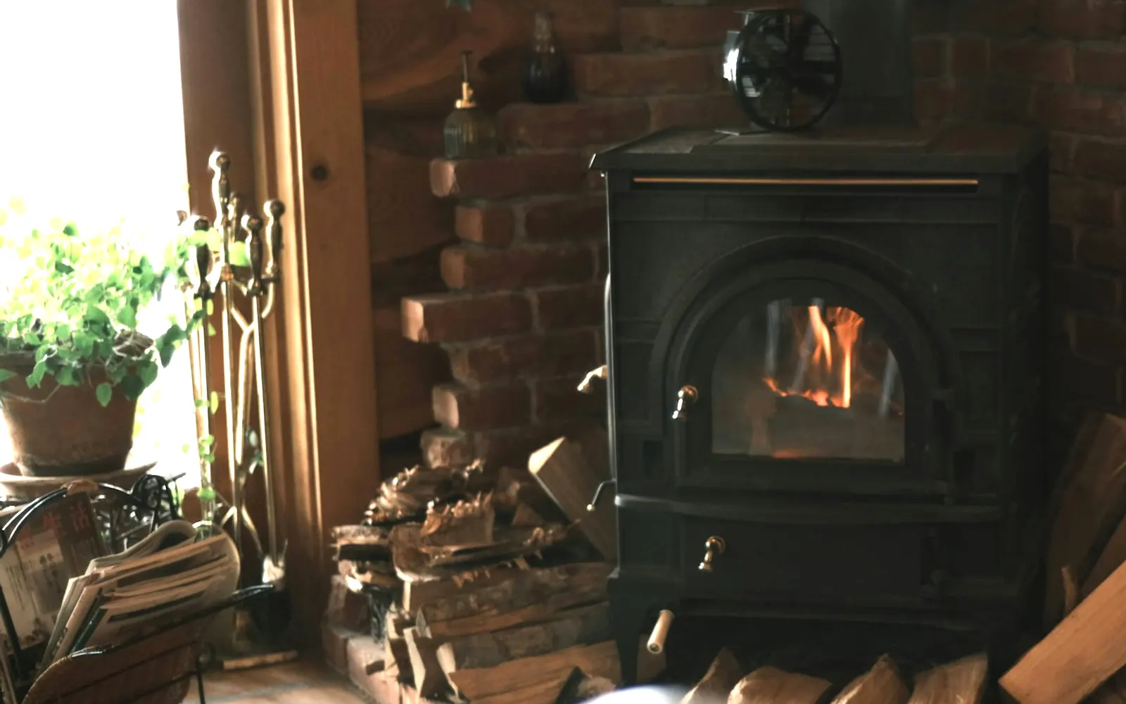 Cast-iron wood stove burning on a brick hearth with split firewood stacked beside it