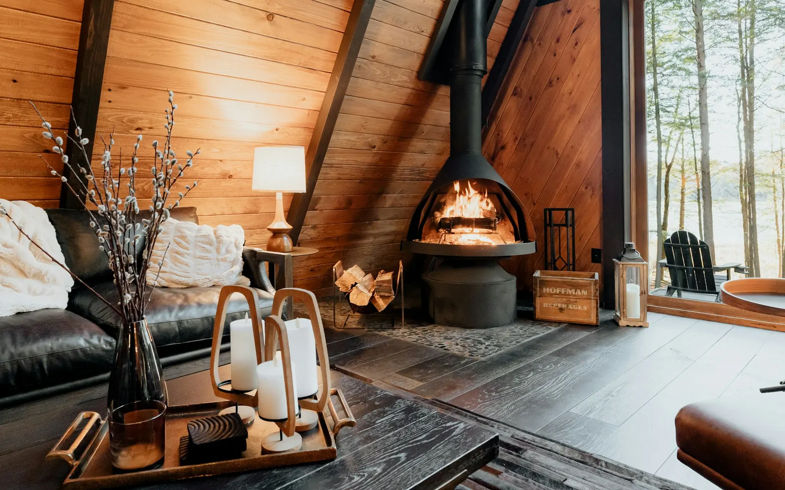 Freestanding round fireplace burning in a wood-paneled A-frame cabin with big windows to the trees
