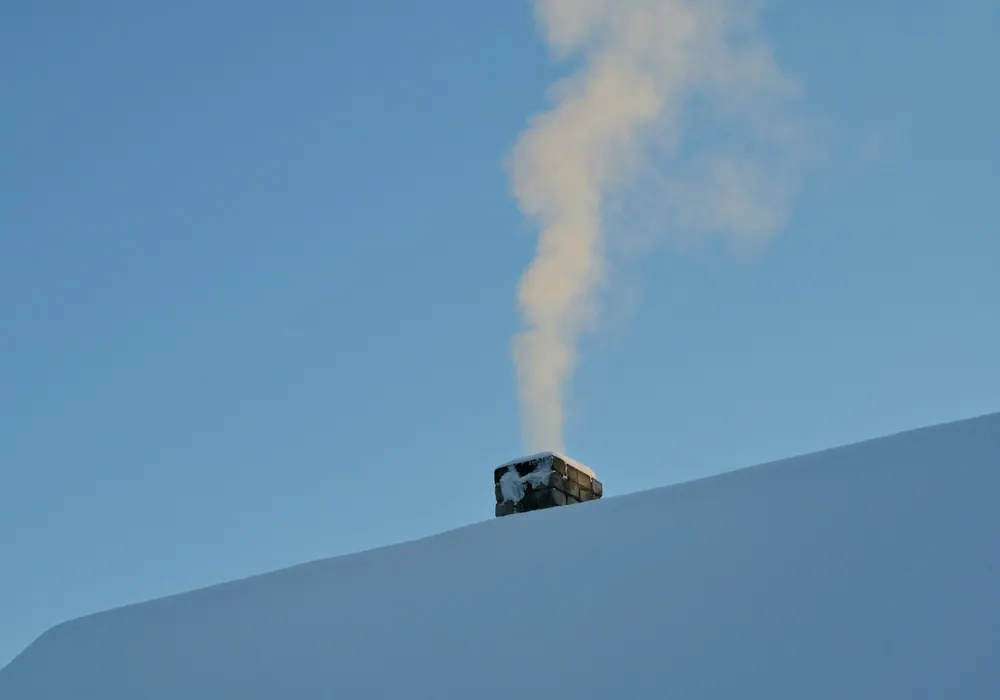 Chimney cap on a snow-covered roof with smoke rising against a blue sky
