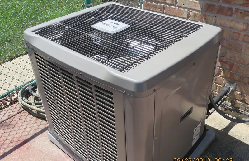 A gray central air conditioning condensing unit on a concrete pad next to a brick house and chain-link fence