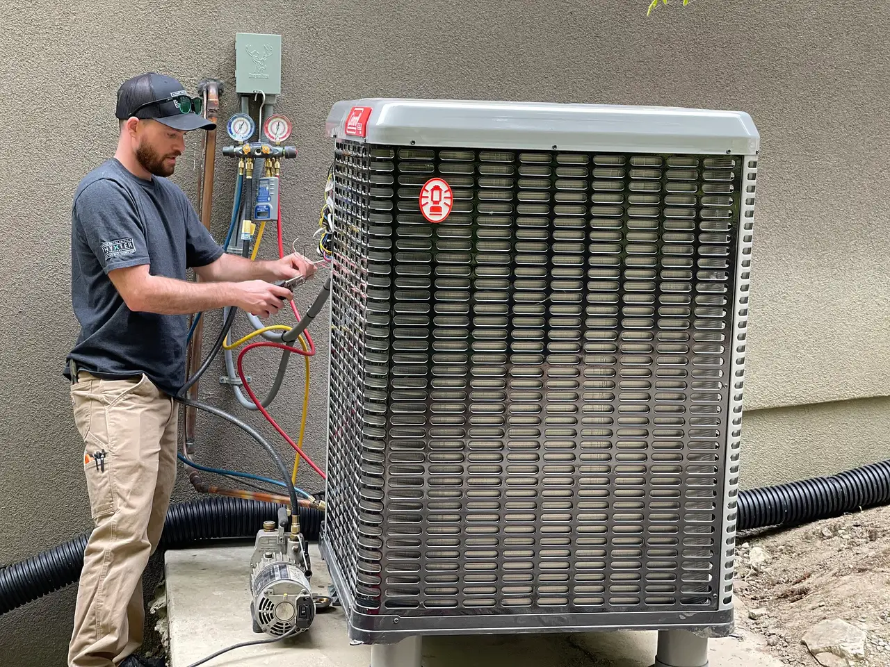 An HVAC technician with refrigerant gauges and hoses connected to an outdoor condensing unit beside a house