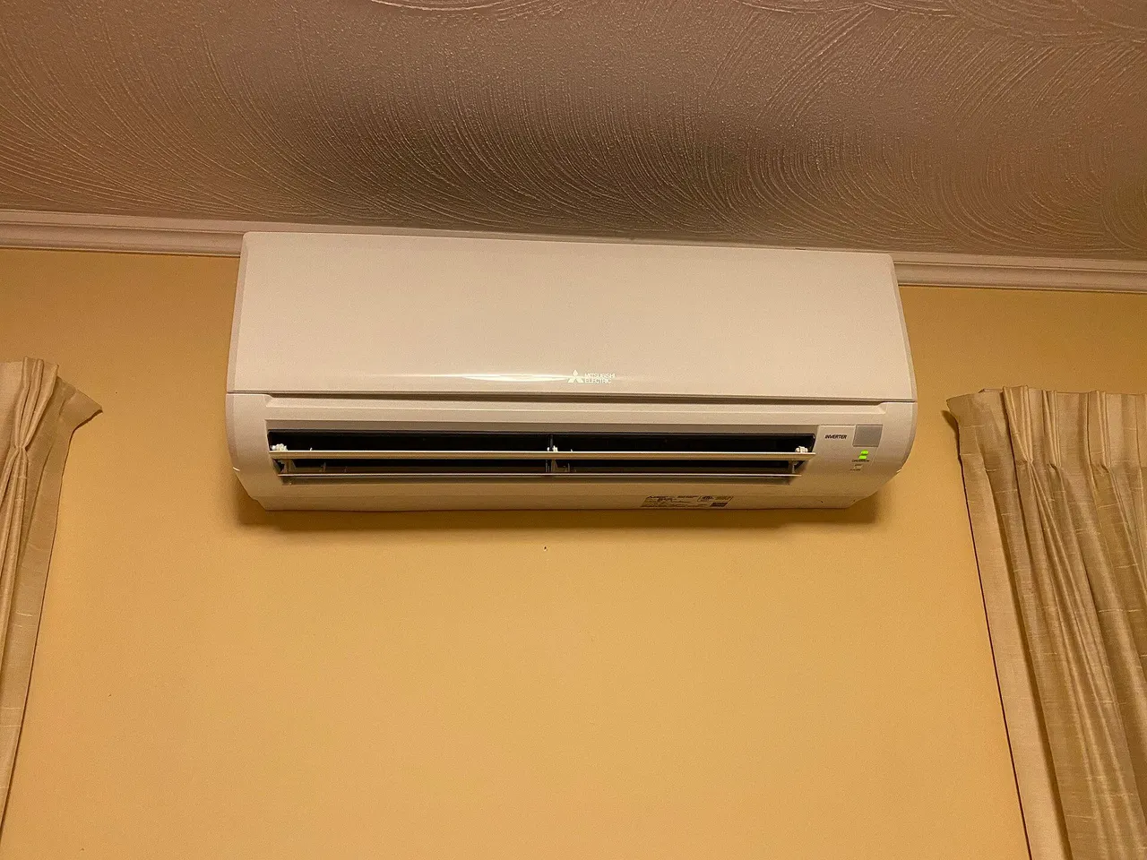 A white Mitsubishi ductless indoor unit mounted high on a yellow wall between curtains