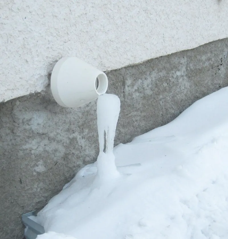 A white PVC furnace exhaust vent on a house wall with a long icicle of frozen condensate running down into snow