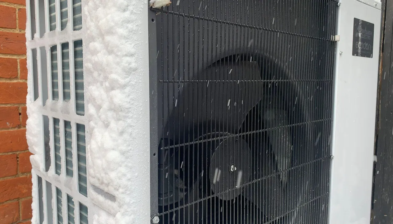 An air-source heat pump outdoor unit with its fan grille against a wall while snow falls around it