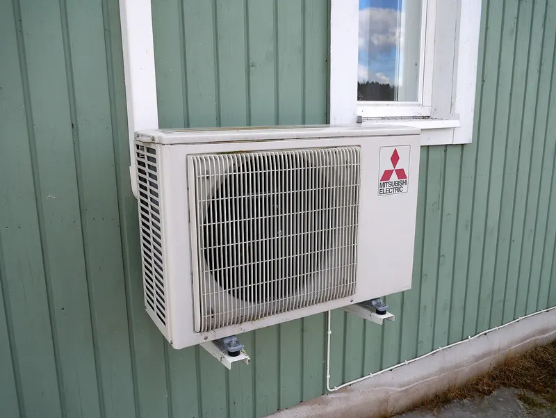 A Mitsubishi Electric mini-split outdoor unit mounted on a wall bracket on green siding below a window
