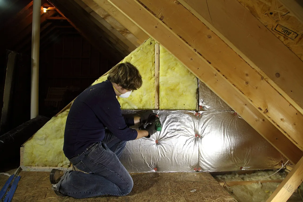A man kneeling in an unfinished attic fits insulation between the rafters, with batts and foil-faced insulation around him