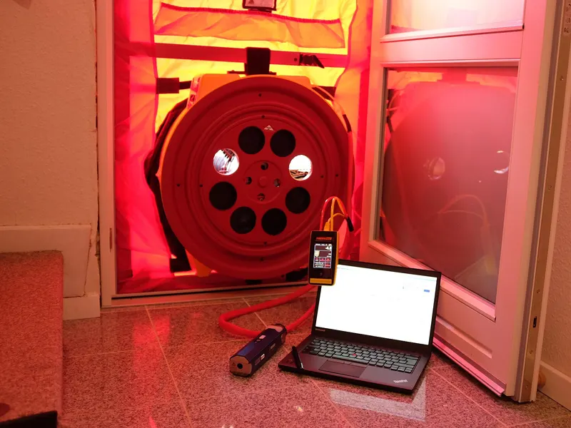 A red blower door fan sealed into a doorway frame, connected to a pressure gauge and a laptop on the floor