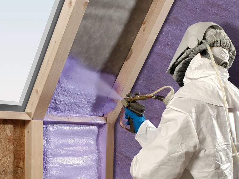 Installer in a protective suit and respirator spraying purple polyurethane foam between wall studs