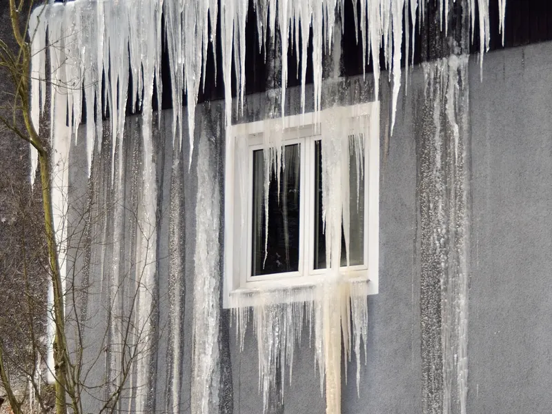Thick icicles hanging from a roof edge down over a window, with meltwater frozen down the wall