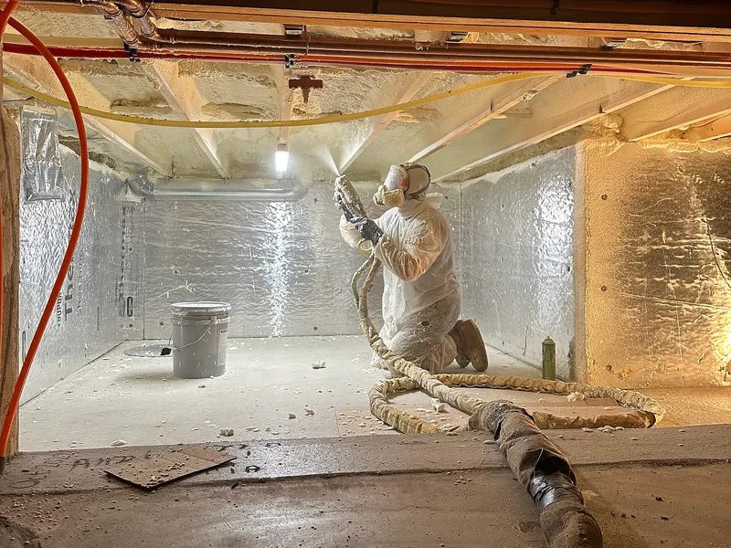 A worker in a white protective suit and respirator sprays foam insulation on the walls of a crawl space, with a hose running across the floor