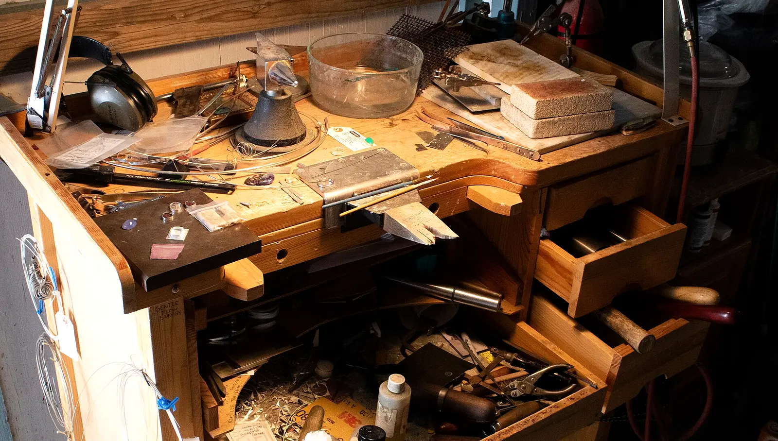 A goldsmith’s wooden bench pin workbench under a lamp, with a ring mandrel, torch blocks, files, pliers and loose gemstones