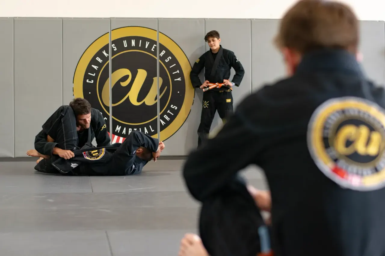 Students in black gis drilling on the mat beneath the gold Clark’s University logo while a coach watches