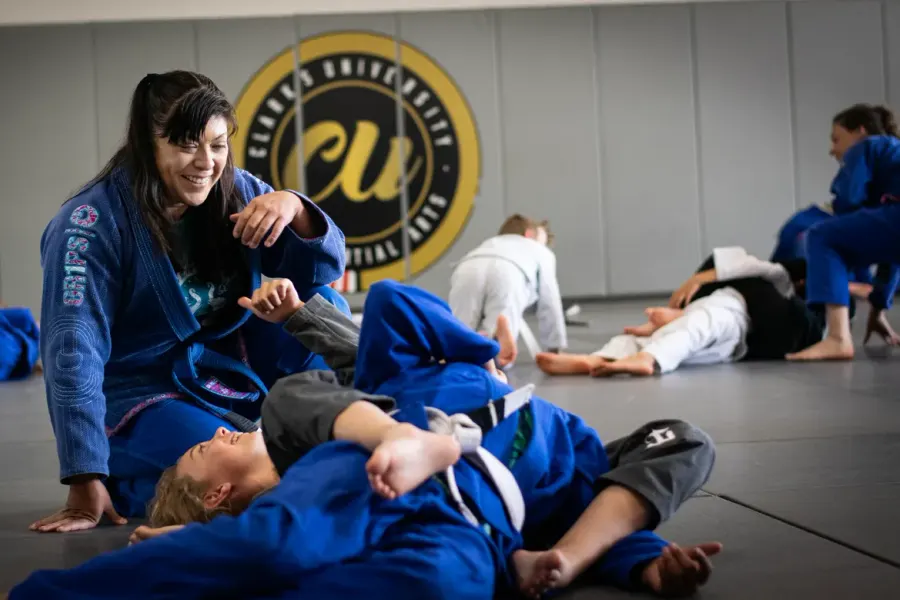 A smiling coach in a blue gi kneels beside two kids practicing on the mat at Clark’s University